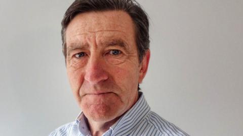 A closeup of Derek Flint, a middle-aged man with dark hair who is wearing a striped shirt and looking sternly at the camera.