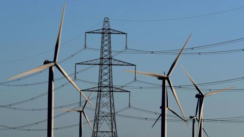 A group of five wind turbine, in shadow against a blue sky, stand in front of a large electricity pylon with cables stretching to either side.