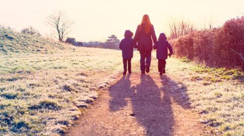 A woman with her back to the camera walks on a path, through a field, holding the hands of two children. They are all wearing trousers and dark winter coats and there is frost on the ground.