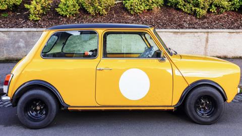 A yellow Mini, side on, in a street, with a path in front, a wall, and wood chippings and foliage. 