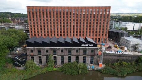 A red brick multi-storey building is surrounded by a building site. There is a smaller building in front that has a sign that reads Good Yards. With brown water in front of that.