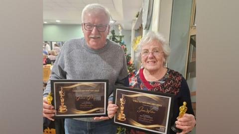 A man and a woman, who are both retired and work as volunteers, stand in a hospital atrium holding framed "Voluntary Oscars" certificates and miniature gold-coloured, Oscar-style trophies. Graham stands on the left in a grey jumper and jeans, while Jean wears a sparkly jumper.