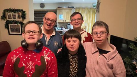 A family of five brothers and sisters are standing together and smiling for a photograph. There are two women and a man on the front row and two men on the back row. One of the men on the front row is wearing a Christmas jumper with a deer's antlers just visible.