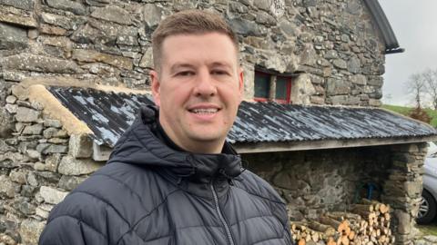 A photo of Jonny Walker who has short brown hair, and is wearing a black puffer coat standing outside in front of wood blocks on the side of a farm building. 