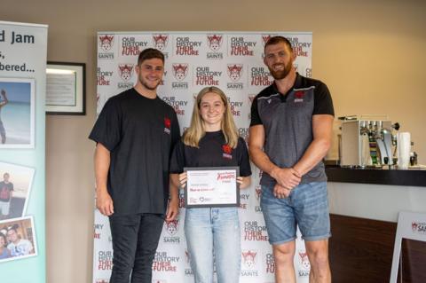 A woman with blonde hair standing between two men and holding a certificate 