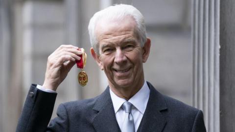 Man with grey hair, smiling at camera, in dark grey suit and white shirt and silver tie, holding up a medal with a gold and red ribbon.