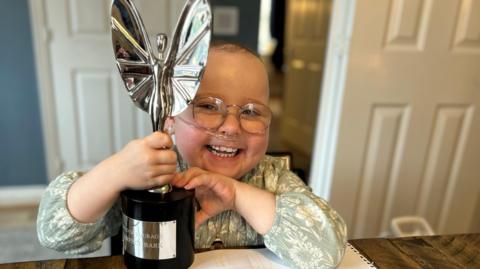 Florrie Bark holding Pride of Britain award. She is wearing glasses and grinning at the camera.