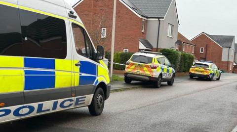 A police van and two police cars are parked in a residential street. Police cordon tape is across one of the driveways.