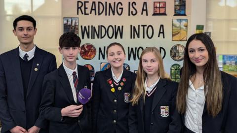 Five teenagers stand in a line wearing school uniform. There are two boys on the left and then three girls to the right. They are wearing dark blue blazers and white shirts, some are wearing ties. One of the boys is holding a purple BBC Radio WM microphone.