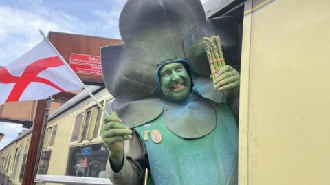 A man in an asparagus costume holding a bundle of asparagus in one hand and the flag of England in the other whilst hanging out of a train