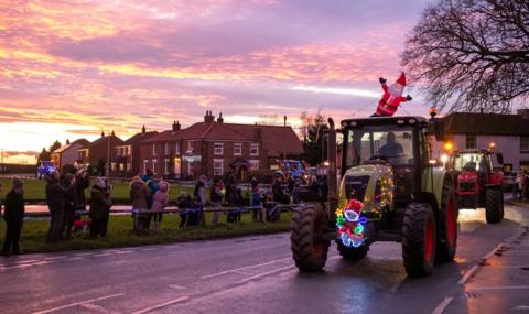 A line of tractors in procession through a village, there is a green tractor with an inflatable Santa on top. People line the streets to watch the tractors come by. There is a pretty pink sky at sunset.