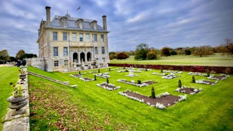 The Italian inspired country mansion of Kingston Lacy sits in formal gardens with a grassy sloping bank. Cloudy skies sit behind it as well as parkland with trees.