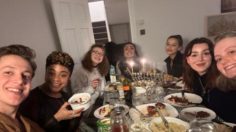 In this photograph Rebecca is pictured with six of her friends around a dinner table, enjoying a feast. In the centre of a table is a menorah, a candelabra lit during the festival. 