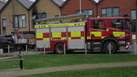 A fire engine in front of a row of houses in Wild Ash Croft, Sittingbourne. One window is damaged by the fire in a house behind the fire engine.