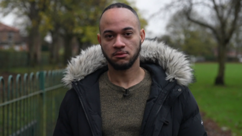 26-year-old Derai with a serious facial expression. He is wearing a brown woollen jumper and a dark coat with an artificial fur collar.