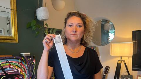 Isabelle Pepin, a woman with blonde hair tied back, holding a copy of the fine issued to her by BCP Council. It looks like a long receipt you would get from a supermarket. She is standing in her living room. In the background is a lamp and a round mirror on the wall