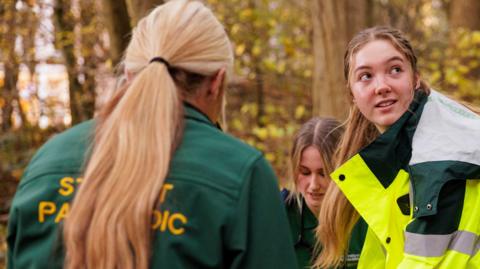 Three trainee paramedics, all female. One to the right has a long ponytail, wearing a uniform and high-vis jacket, she is looking to the left, one behind her is looking down and a third, with a long blond ponytail, has their back to the camera. They are in a wood, with trees behind them. 