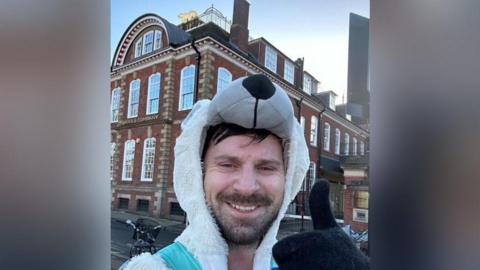 Alex is smiling at the camera with his thumb up, taking a selfie. He is wearing a white polar bear onesie, with the hood up, which is the bear's head. In the background is a building with a polar bear sculpture lit up on its roof