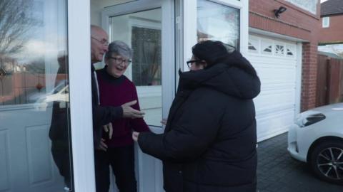 Mark and Elisabeth Bryant stand in their door and welcome Yasmina inside.