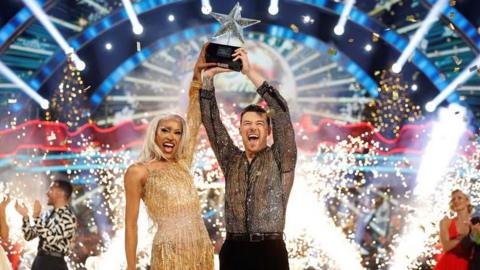 Drag artist Tayce in gold dress lifts star-shaped trophy with dance partner Kai Widdrington on Strictly Come Dancing Christmas special as bright lights sparkle in the background