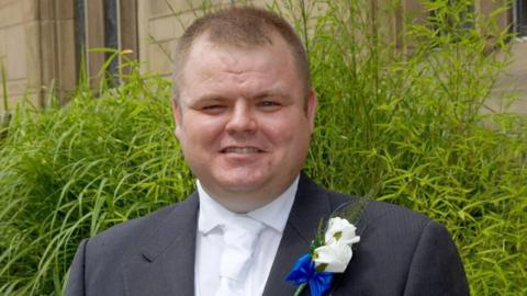 PC Neil Doyle, who has short blonde hair, smiles at the camera wearing a grey wedding suit over a white shirt, and has a white flower pinned to his lapel