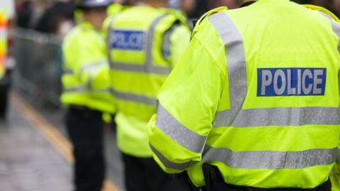 Three police officers. Their backs are facing the camera and they are wearing yellow fluorescent jackets. On the back of the jacket it says POLICE in blue and silver writing.