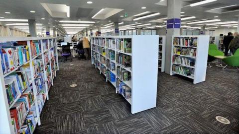 Rows of books on white shelves in a library in Wakefield. There is a grey patterned carpet and people on a computer in the background.