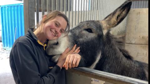 A woman in a black fleece with yellow trim is leaning into a donkey stood in its paddock. Her hand is stroking beneath the donkey’s mouth. She has dark hair tied back. The donkey has a white nose and a dark grey face and body. The barn door can be seen in the background.