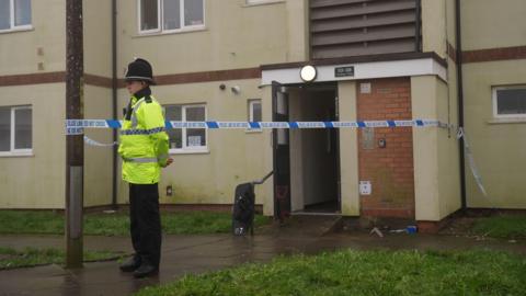 A police officer in a yellow high visibility jacket stands by a police cordon which is surrounding the entrance to a block of flats. There is police riot equipment visible by a doorway.