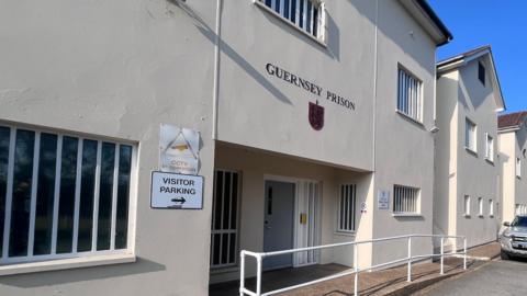 The outside of Guernsey Prison. It is a cream-coloured building with a shield and the words GUERNSEY PRISON above the main entrance. There are two other signs, one reading VISITOR PARKING and the other CCTV IN OPERATION to the right of the entrance. A ramp and white railings lead to the grey front door. The windows feature many struts from top to bottom.