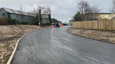 A freshly paved road with exposed earth on either side, a wooden fence on the right side of it, and a corrugated industrial building on the left. Further down the road in the distance are orange traffic cones and a group of people.