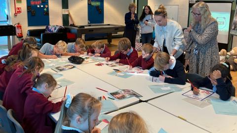 A class room of children sat around a large table. There are a few teachers stood around the table.