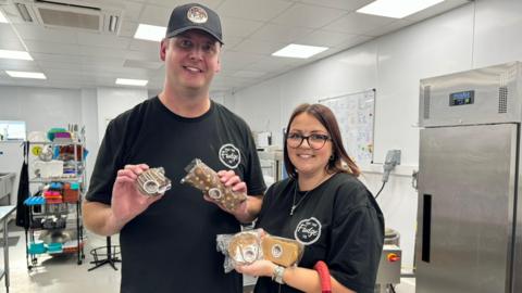 Pete and Claire Brennan in their factory kitchen. They are both wearing black T-shirts featuring their company logo. Pete is also wearing a black baseball cap. Claire has dark hair and is wearing glasses. The couple are both holding a selection of fudge and chocolate products.