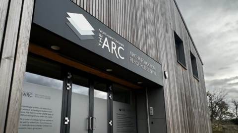 A wooden building with glass doors with frosted glass. A black sign above the door reads "Northamptonshire Archaeological Resource Centre".