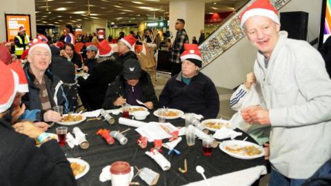 A group of men are sitting around a table inside Birmingham New Street station. They are wearing red Santa hats and eating food from paper plates. 