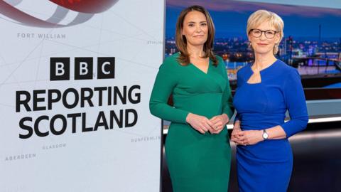 The Reporting Scotland studio with presenters Laura Miller and Sally Magnusson standing next to a the logo for the programme on a white background