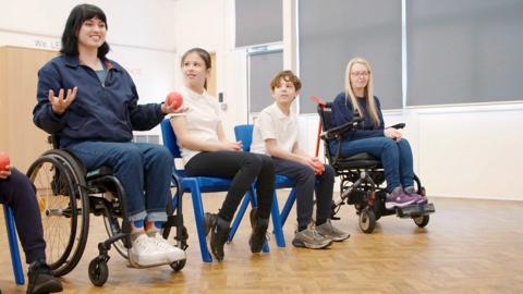 A presenter in a wheelchair in a school sports hall demonstrates boccia with children and teachers watching.