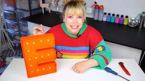 A lady smiles at the camera whilst sitting at a desk. On the desk is a large light up letter E, a screwdriver and a scalpel.