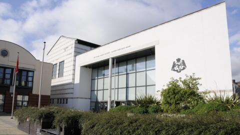 The frontage of a modern courthouse building, with a large glass entrance and the Isle of Man government crest on its white walls. There are a collection of bushes in the foreground.