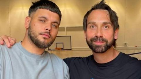 Two bearded men side by side. One wearing a grey t-shirt, one wearing a black t-shirt