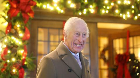 King Charles III. He is pictured outside an entrance which is decorated for Christmas. Fairylights, red ribbon and wreaths can be seen in the distance, although they are out of focus in the frame. The King is wearing a light brown coat, a white shirt and a light pink tie. He has short white hair and blue eyes. He is looking away from the lens, smiling. 