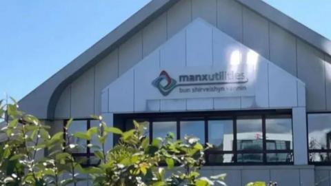 The exterior of the Manx Utilities building, which is grey with a pointed roof and a colourful sign with the firm's name. There are green bushes in the foreground.