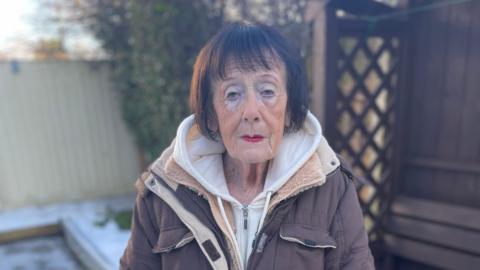 An elderly woman, wearing makeup and a brown coat, with short brown hair, stands outside in her snowy garden, looking at the camera 