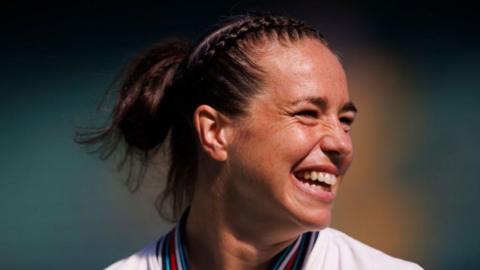 A woman is looking to the right, smiling. She has her hair plaited into a bun.