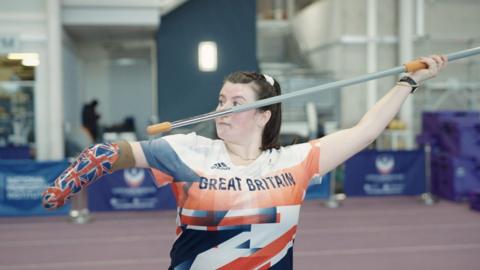 Paralympic athlete Hollie Arnold holds a javelin in an athletics arena wearing a Great Britain team shirt.