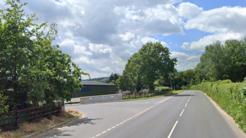 Google image of the entrance to Beacon Park near Dartington. Green hedges and trees line either side of the road outside of the entrance. A large white sign states Beacon Park. 