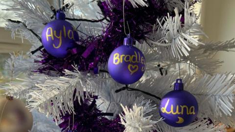 A close up of a white Christmas tree. There is purple tinsel on the tree and string lights. There are three purple ornaments going down the tree whith gold writing. Left to right -  the ornaments have the names Ayla, Bradley and Luna