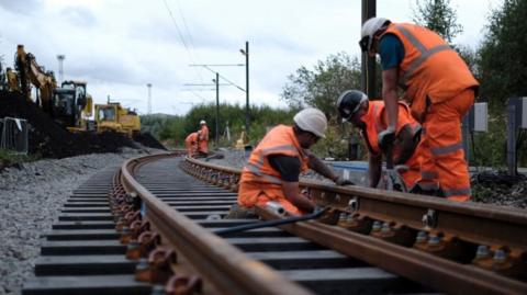 Three workers are on the track in the foreground. Others, also wearing orange hi-vis jackets and protective headwear, are in the distance.