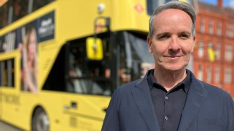 Sacha Lord, wearing a dark blue blazer over a black shirt, stands in front of a bright yellow Bee Network bus 