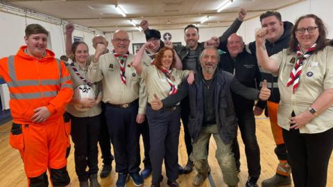 A group of 11 people stand in a scout hut some with their arms raised in celebration. A number are wearing scouts uniform.  